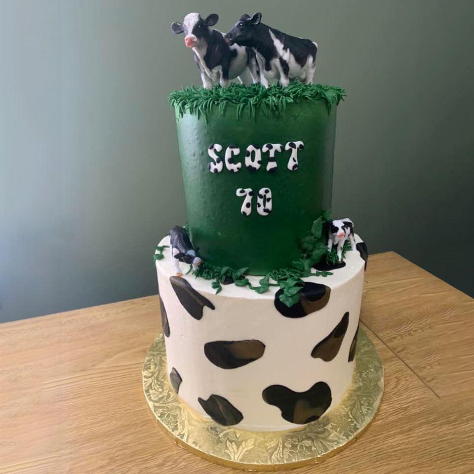 Dark green cake with cow cake toppers that reads "Scott 70" over top of a cake with a cow patt